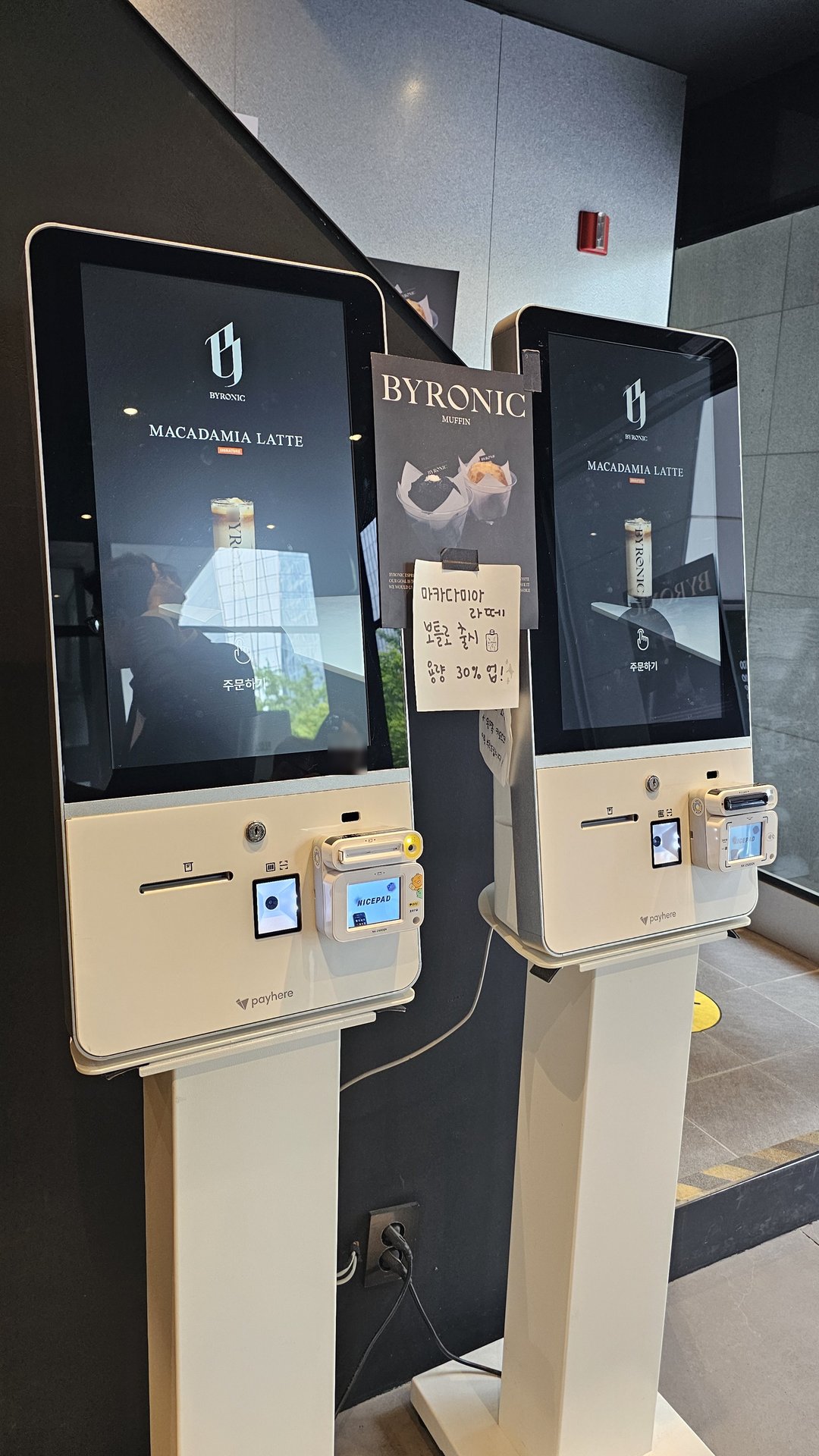 Self-order kiosk screens at Byronic Espresso Yeouido showing Macadamia Latte menu and muffin promotional leaflet