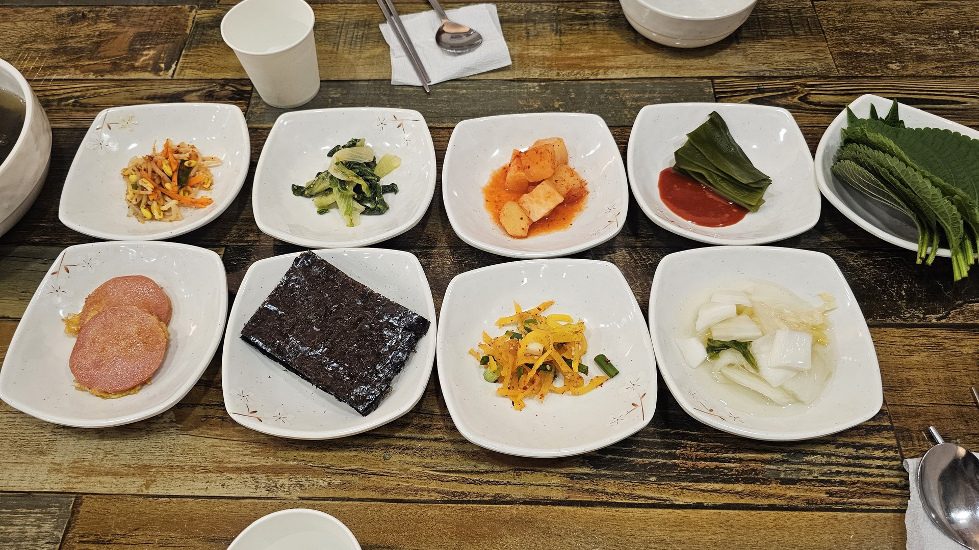 Eight-piece banchan spread including egg roll, spam, seaweed, bean sprouts, kimchi, perilla, and cucumber