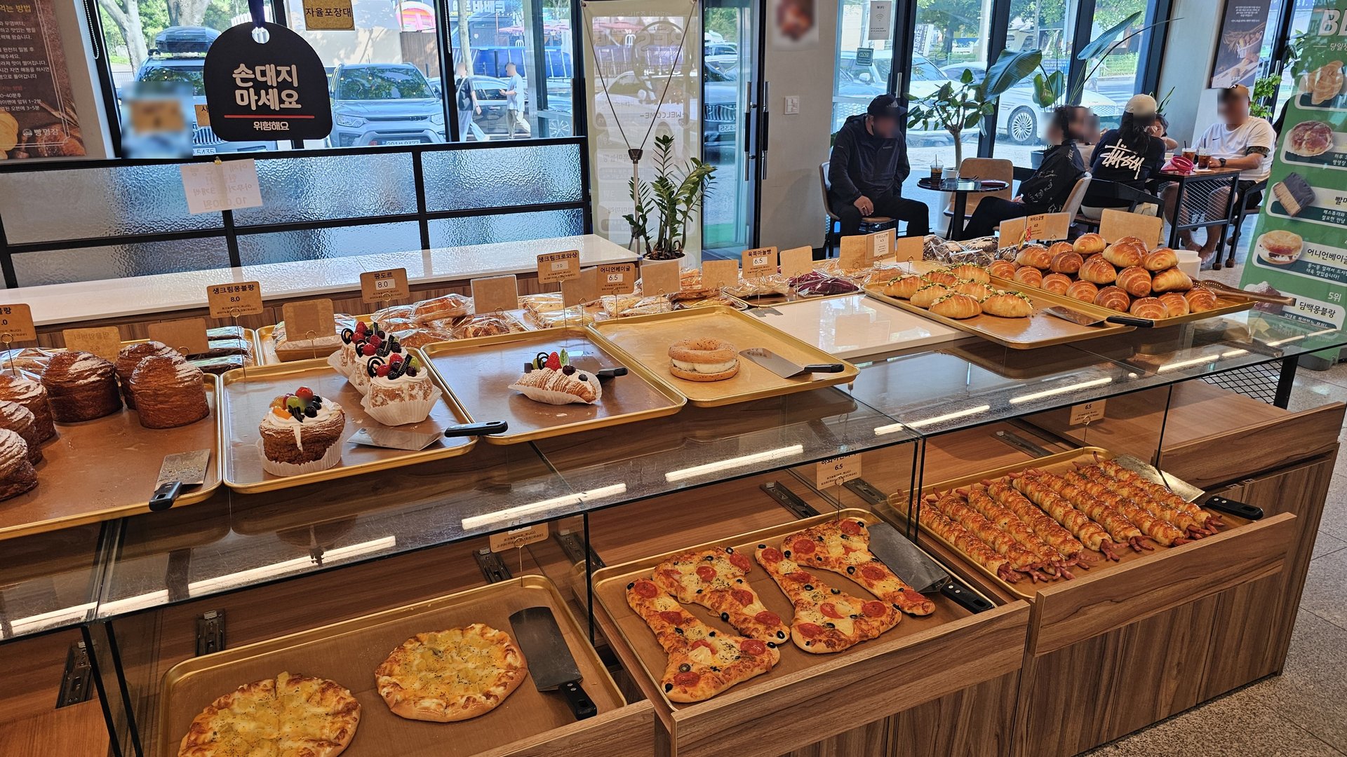 Pizza bread, cakes, donuts, and croissants in display counter with customers browsing at Mochang-po Bread Cafe