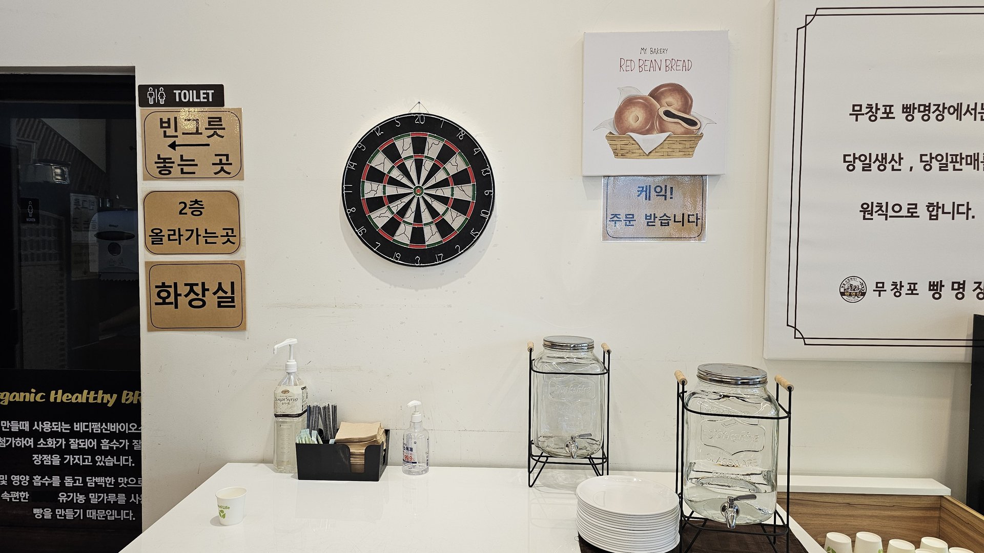 Interior wall with dartboard, cake order sign, same-day baking policy notice, and water dispenser at Mochang-po Bread Cafe