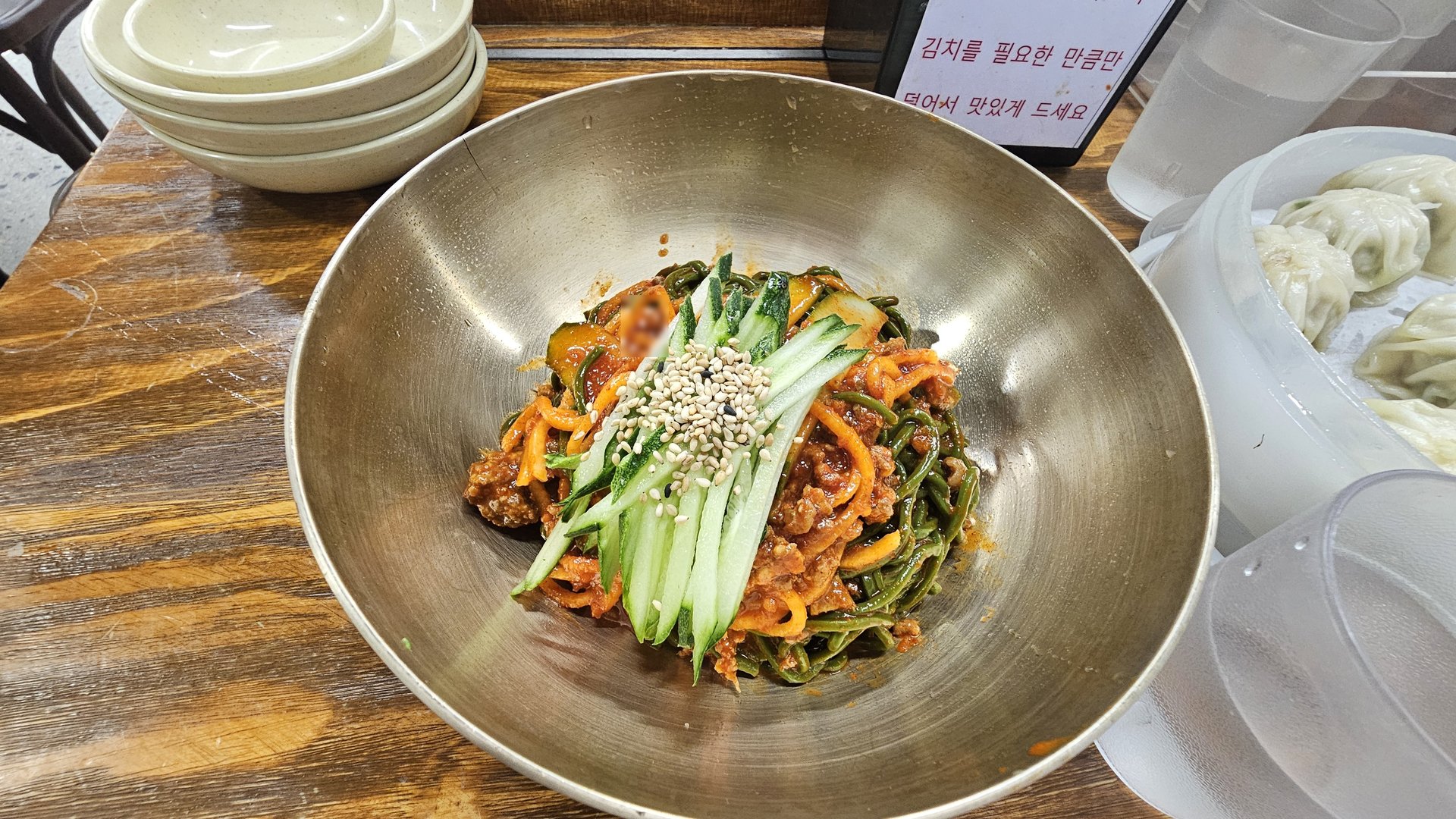 Bibim guksu spicy cold noodles in large stainless steel bowl topped with cucumber perilla and sesame