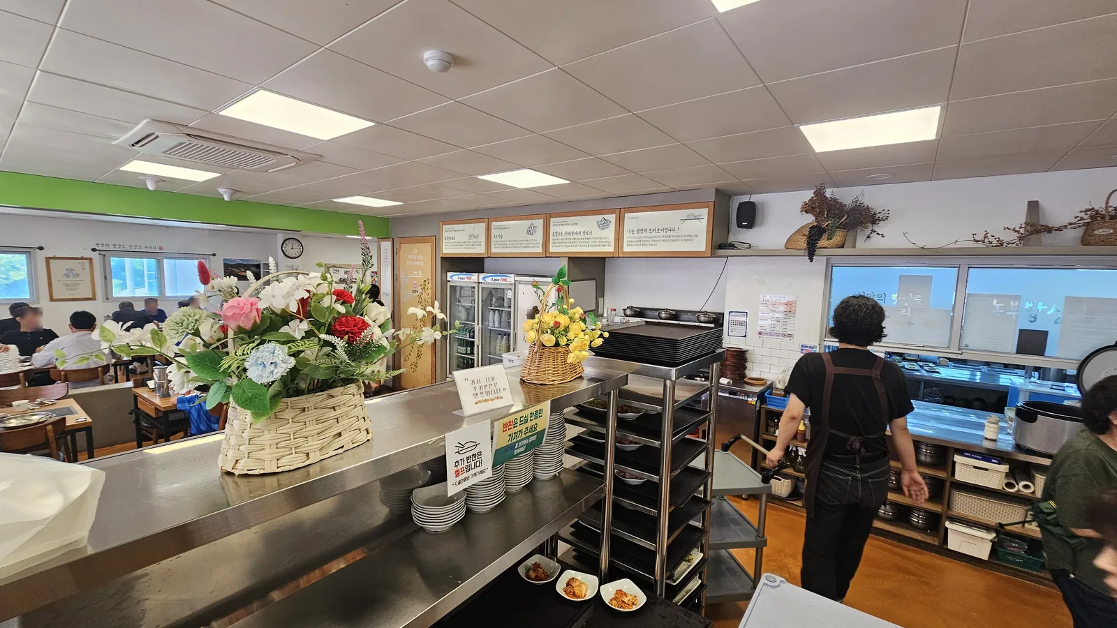 Self-service banchan bar and kitchen counter at Nongbu Bapsang with staff preparing dishes and floral decoration