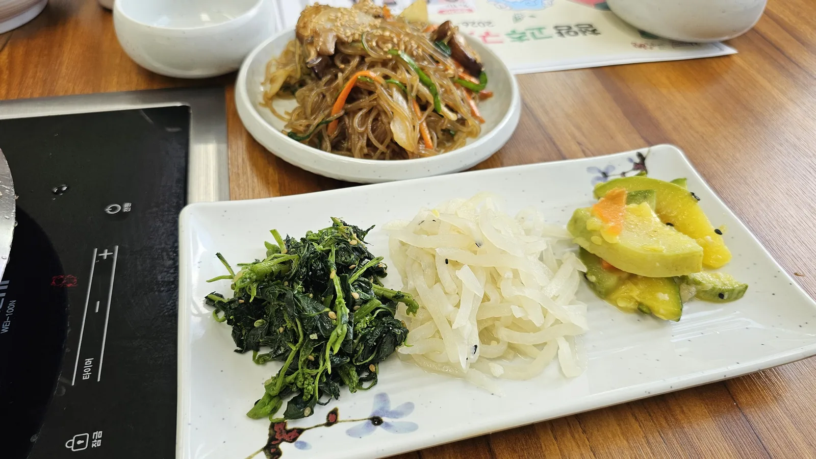 Nongbu Bapsang banchan plate with chive namul, radish strips, and cucumber salad alongside japchae glass noodles