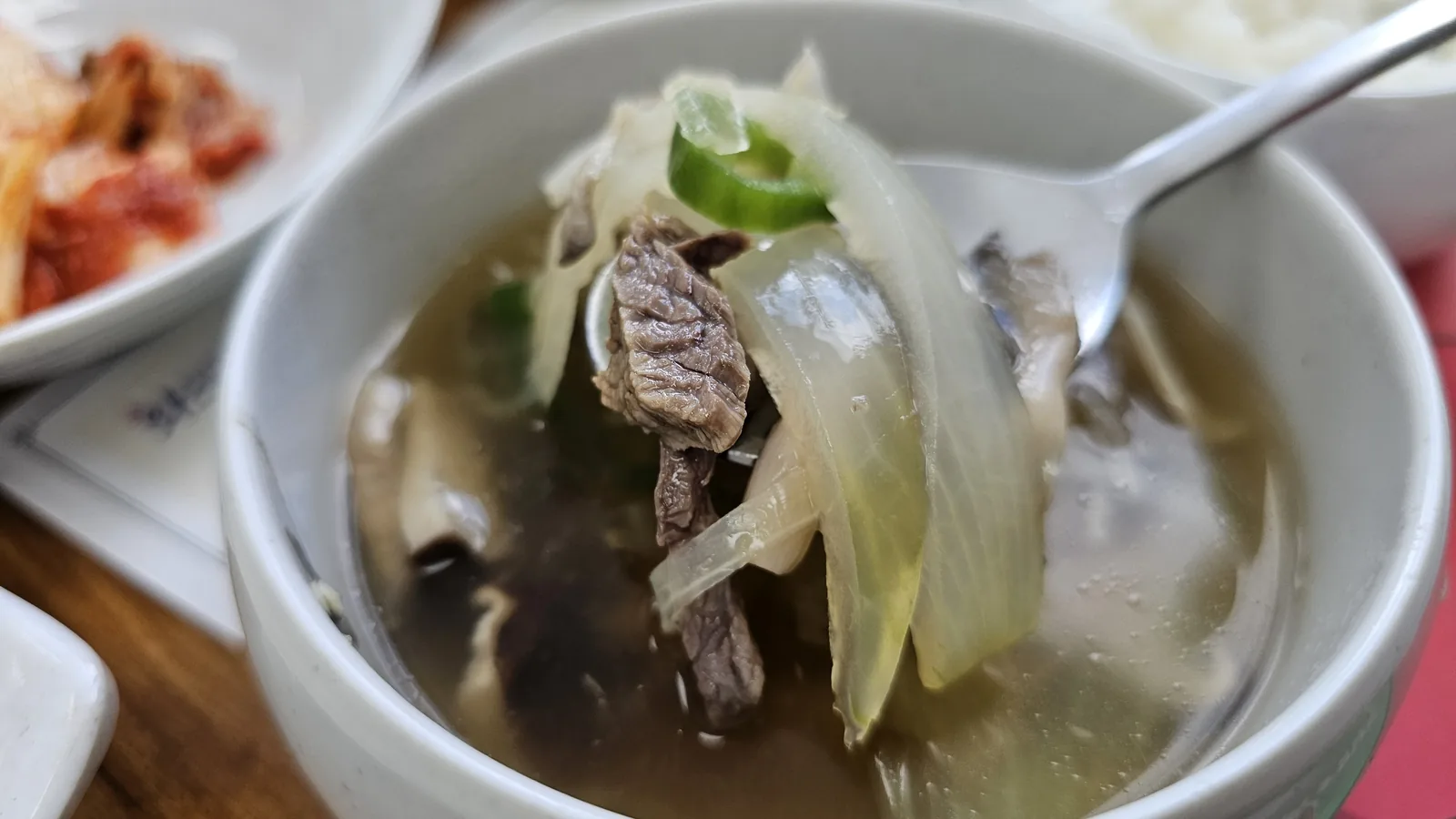 Clear hot pot broth close-up at Nongbu Bapsang showing thin-sliced meat, onion, green chili, and mushrooms in light soup